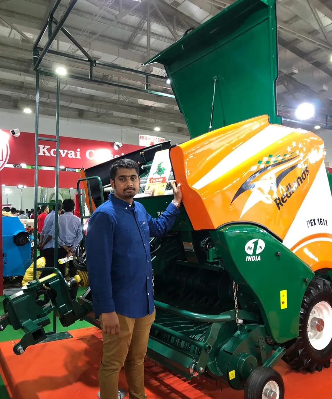 Adhith beside a green and orange agricultural machine at a trade show